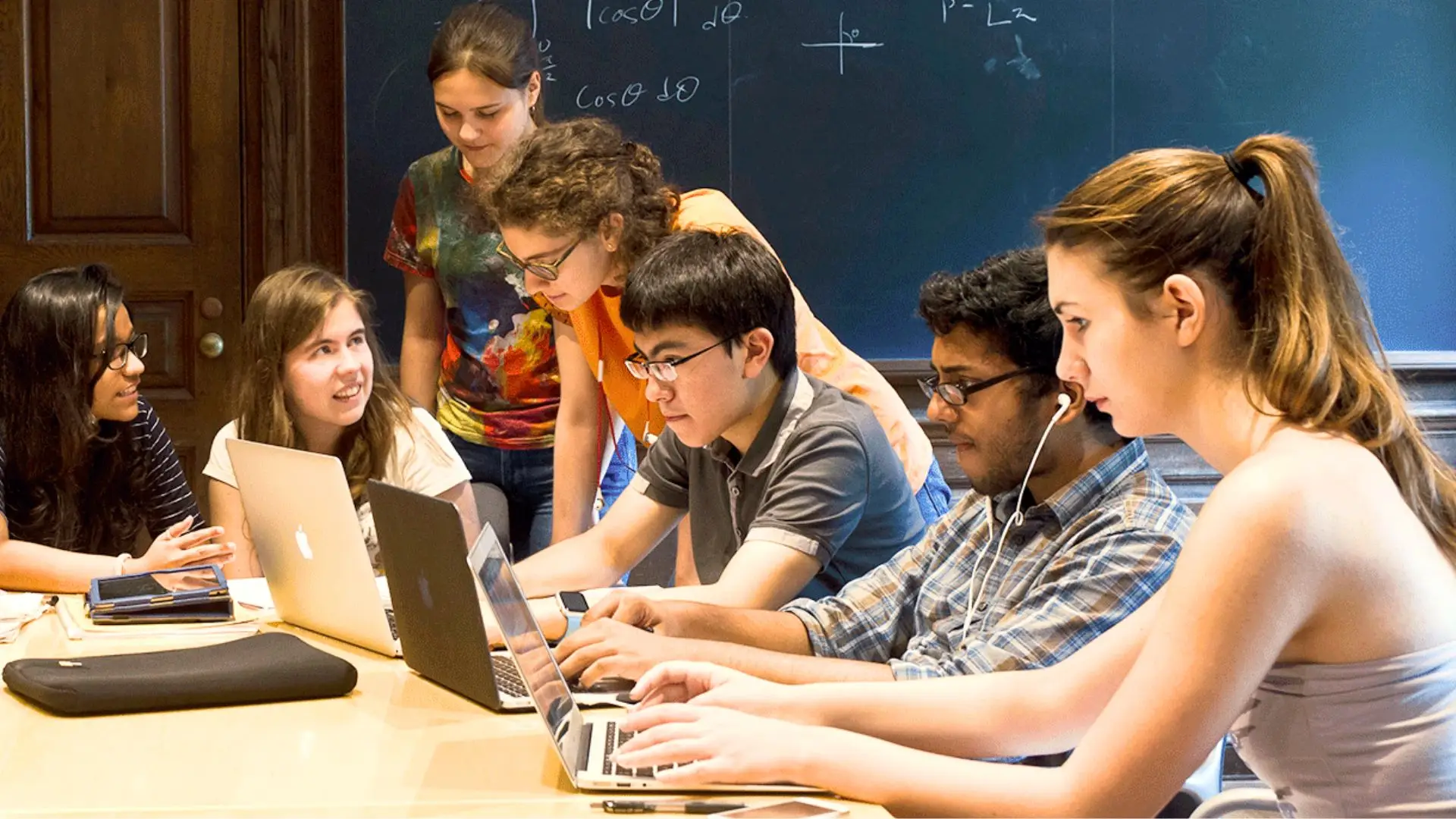 Diverse group of students working collaboratively on laptops at a wooden table in a classroom with mathematical equations written on a chalkboard behind them