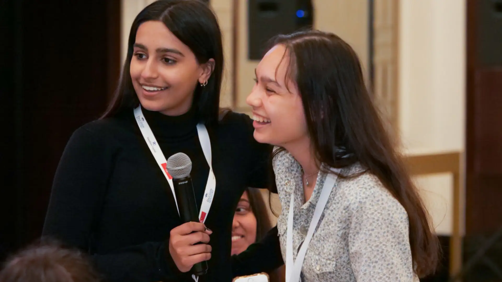 Two confident young women at a leadership conference, one holding a microphone, both smiling and engaged with their peers