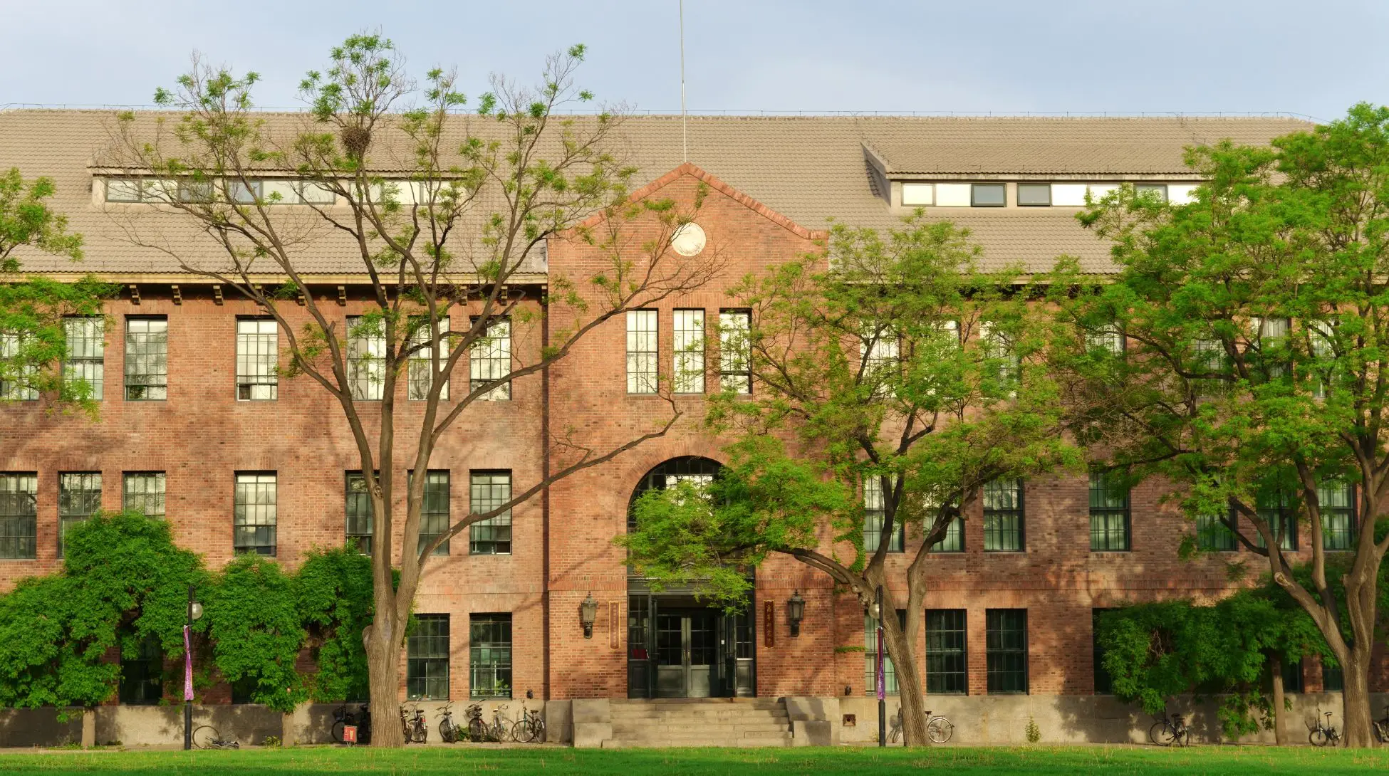 University campus red-brick building with green trees, central entrance, and bicycles parked in front
