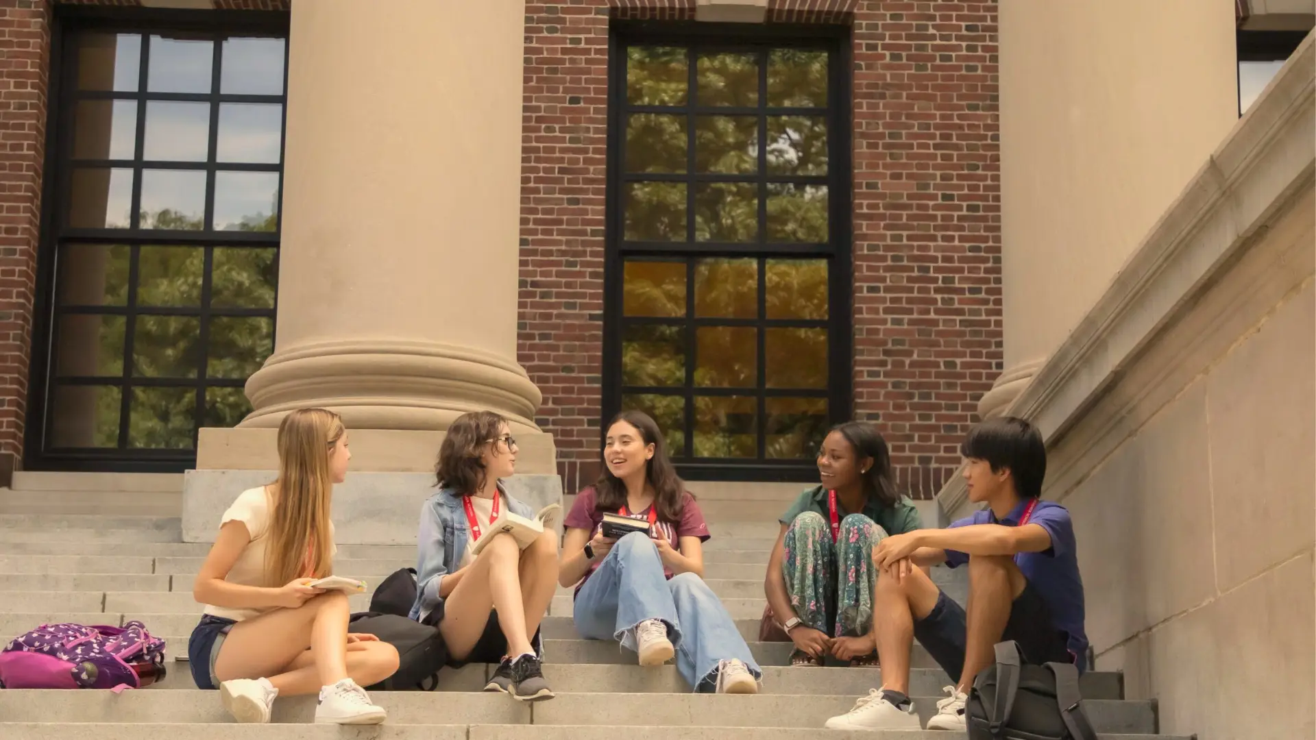 Five diverse students sitting on university library steps chatting and laughing together with books and backpacks